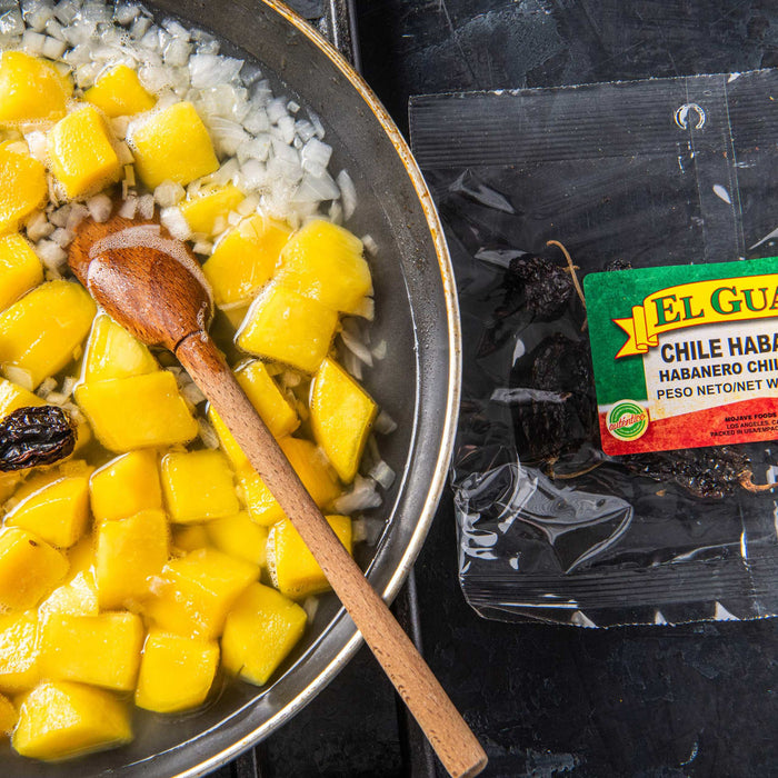A close-up of a pan of diced mango and onions with a wooden spoon, next to a bag of El Guajolote Habanero Chiles.