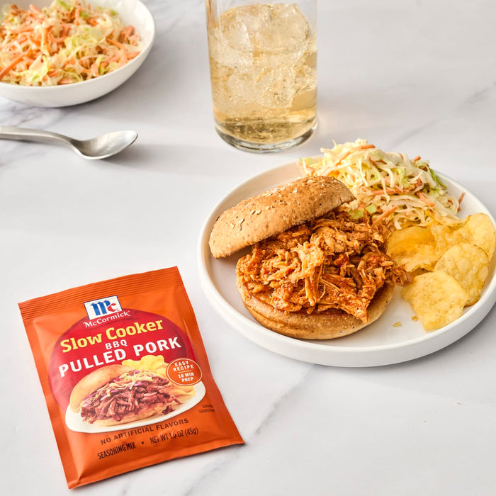 A packet of McCormick Slow Cookers Pulled Pork seasoning is displayed next to a pulled pork sandwich, coleslaw, and potato chips.