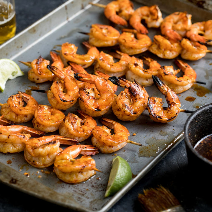 Grilled shrimp skewers, seasoned with Mccormick Grill Mates Chipotle &amp; Roasted Garlic Seasoning, are arranged on a metal baking sheet with lime wedges and a dipping sauce.