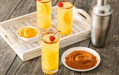 Three orange cocktails with cherries on a wooden tray, accompanied by a cocktail shaker and small bowls of spices.