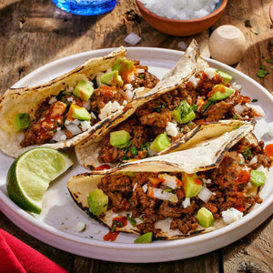 Three flavorful tacos, featuring seasoned meat, diced onions, avocado, and crumbled cheese, are served on a white plate with a lime wedge, suggesting they were prepared using Cholula Original Taco Seas Mix 1 Oz.