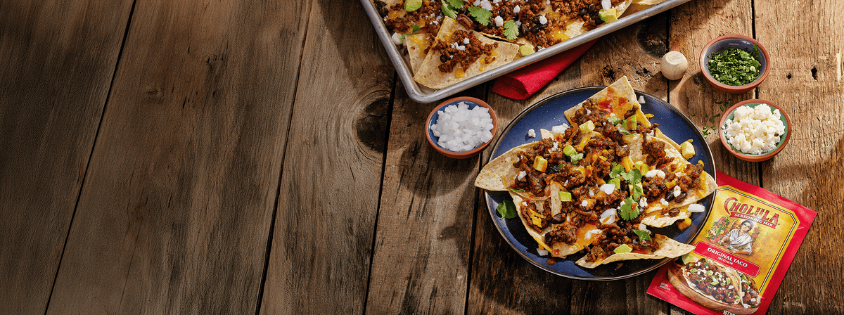 Plated nachos with various toppings on a wooden table, accompanied by small bowls of ingredients and a package of taco seasoning.