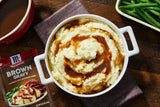 Mashed potatoes with brown gravy in a white bowl on a wooden table, with a packet of McCormick Brown Gravy.