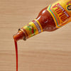 A bottle of Cholula Chipotle Regular Hot Sauce Regular 5 Oz is tilted, with the reddish-brown sauce pouring out in a stream against a light wooden background.