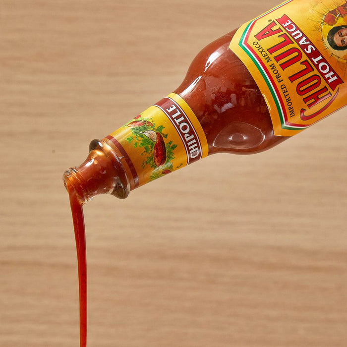 A bottle of Cholula Chipotle Regular Hot Sauce Regular 5 Oz is tilted, with the reddish-brown sauce pouring out in a stream against a light wooden background.