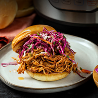 Burger with pulled meat and purple cabbage on a white plate, with a pressure cooker in the background.
