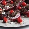 Grilled meat, red onion wedges, and cherry tomatoes, sprinkled with feta cheese and Mccormick Black Pepper Table Grind, are arranged on a light wooden surface.