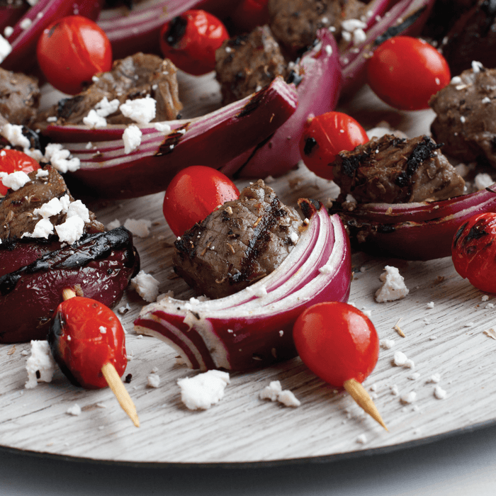 Grilled meat, red onion wedges, and cherry tomatoes, sprinkled with feta cheese and Mccormick Black Pepper Table Grind, are arranged on a light wooden surface.