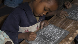 Child writing on a slate with a piece of chalk at a desk.