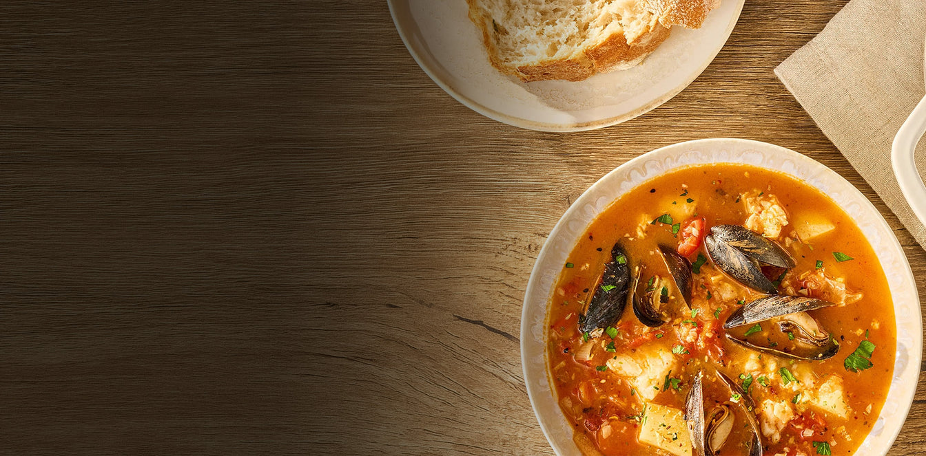 A bowl of seafood and vegetable soup with mussels and tofu on a wooden table, with a plate of bread and a napkin partially visible in the background.