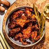 A bowl of glazed chicken wings served with green beans and okra on a wooden board alongside tamarind pods and lemongrass stalks.