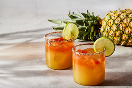 Two tropical cocktails with pineapple and lime garnishes on a wooden surface.