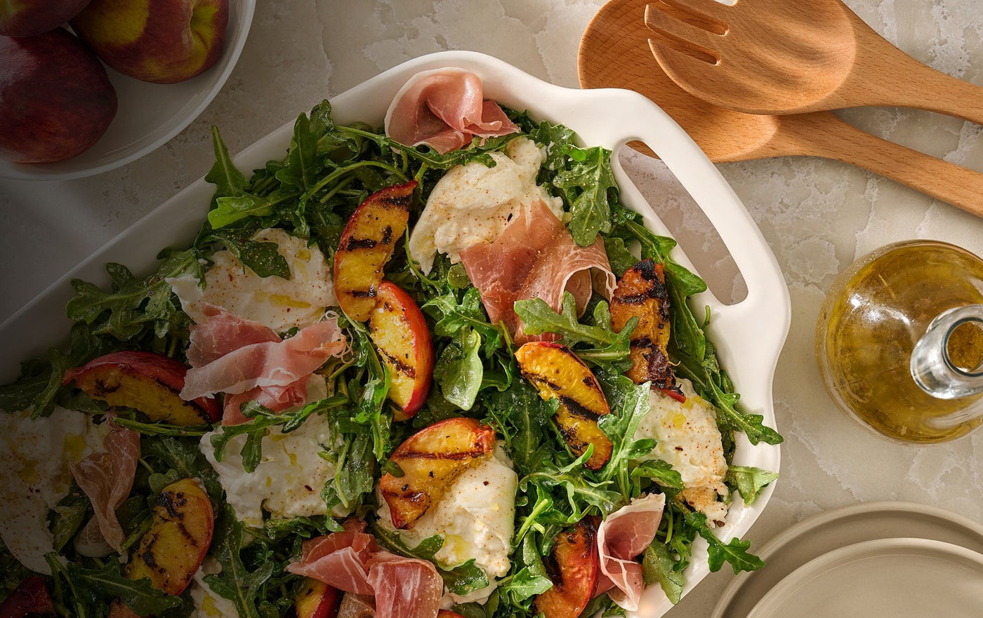 arugula, grilled peaches, and burrata salad on a white marbled counter with a wooden spoon and olive oil carafe next to it.