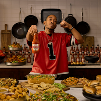 Man in a red sports jersey holding a bottle of Red Hot sauce in a kitchen with food and bottles around.