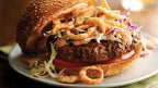 Hamburger with coleslaw, onion rings, and tomato on a sesame seed bun.