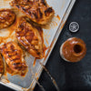 A close-up of a baking sheet with grilled chicken breasts covered in barbecue sauce, a bottle of Texas Stubborn Sauce, and tongs.