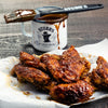 A white mug with the Stubbs brand logo and a brush with barbecue sauce on it, next to a pile of barbecue chicken wings on parchment paper.