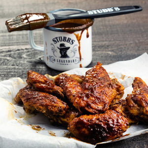 A white mug with the Stubbs brand logo and a brush with barbecue sauce on it, next to a pile of barbecue chicken wings on parchment paper.