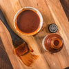 A wooden cutting board with a bowl of barbecue sauce, a bottle of barbecue sauce, and a brush.