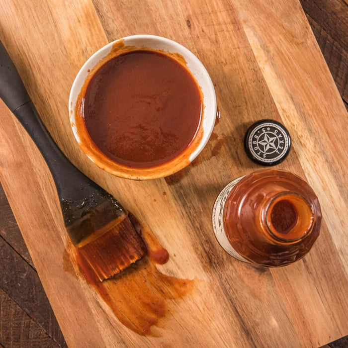 A wooden cutting board with a bowl of barbecue sauce, a bottle of barbecue sauce, and a brush.