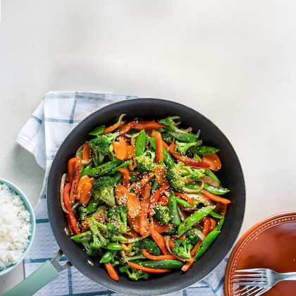 Fried vegetables in a wok with a side of rice on a white table.