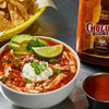 A bowl of chicken chili topped with sour cream, avocado, lime, and green onions, with a bottle of Cholula Hot Sauce and a small bowl of hot sauce in the background.