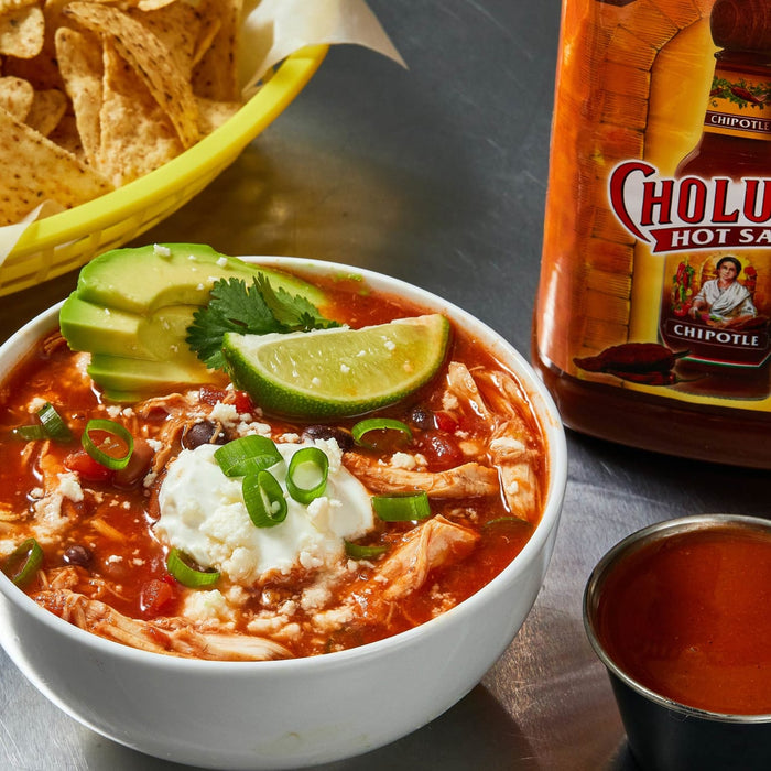 A bowl of chicken chili topped with sour cream, avocado, lime, and green onions, with a bottle of Cholula Hot Sauce and a small bowl of hot sauce in the background.