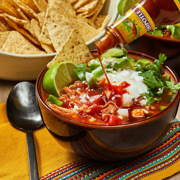 A bowl of chicken tortilla soup with a squeeze of lime, sour cream, avocado, and cilantro, with a bottle of Chipotle hot sauce being poured over the top.