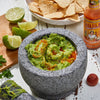A mortar and pestle filled with guacamole and a bottle of Cholula Chili Garlic Hot Sauce.