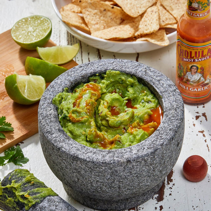 A mortar and pestle filled with guacamole and a bottle of Cholula Chili Garlic Hot Sauce.