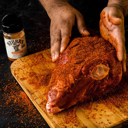 A hand is sprinkling Stubbs Pork Rub on a large piece of meat.