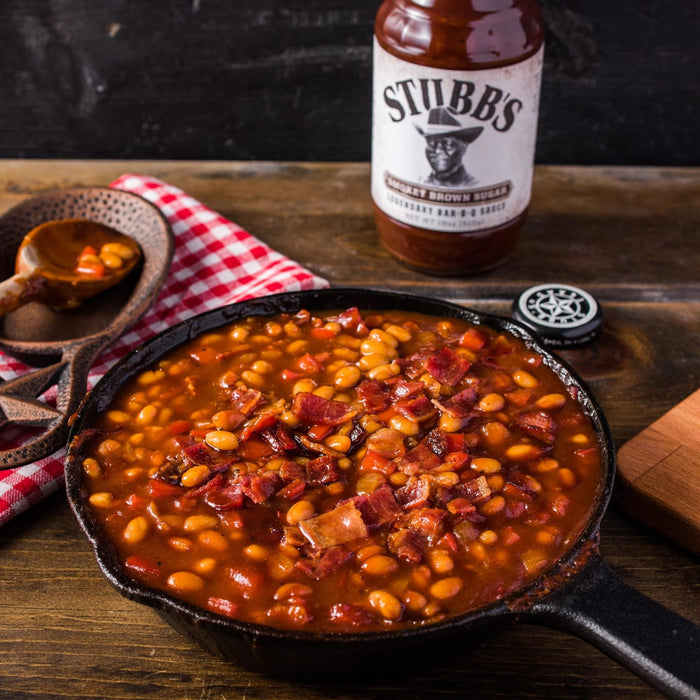 A cast iron skillet filled with baked beans, bacon, and red peppers, with a bottle of Stubbs Smoky Brown Sugar BBQ Sauce in the background.