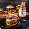 A bottle of Stubbs Simply Sweet Reduced Sugar BBQ Sauce with three burgers topped with avocado and red onion.