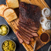 A wooden cutting board with sliced brisket, toasted bread, pickles, jalapenos, onions, and barbecue sauce.