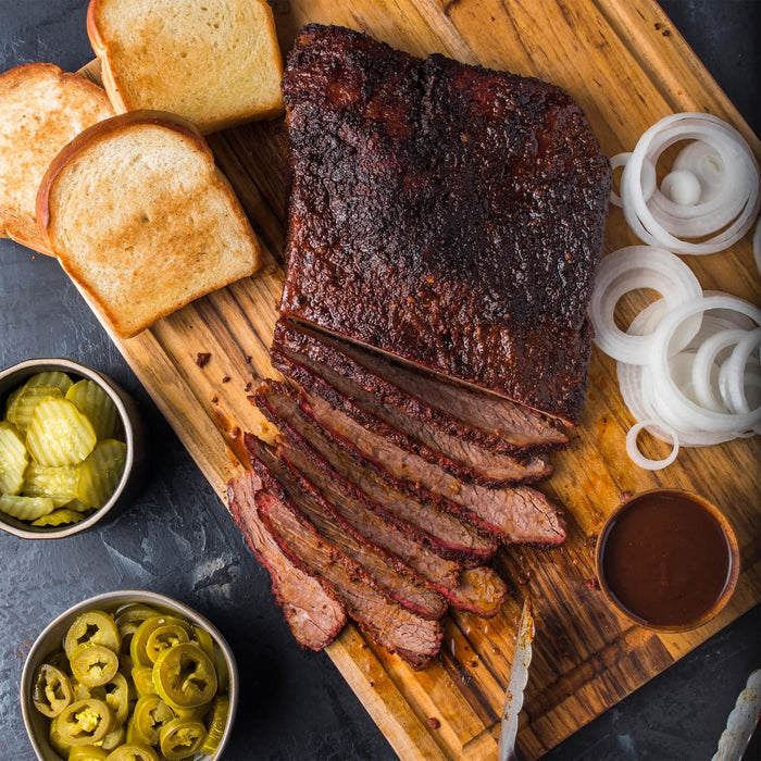 A wooden cutting board with sliced brisket, toasted bread, pickles, jalapenos, onions, and barbecue sauce.