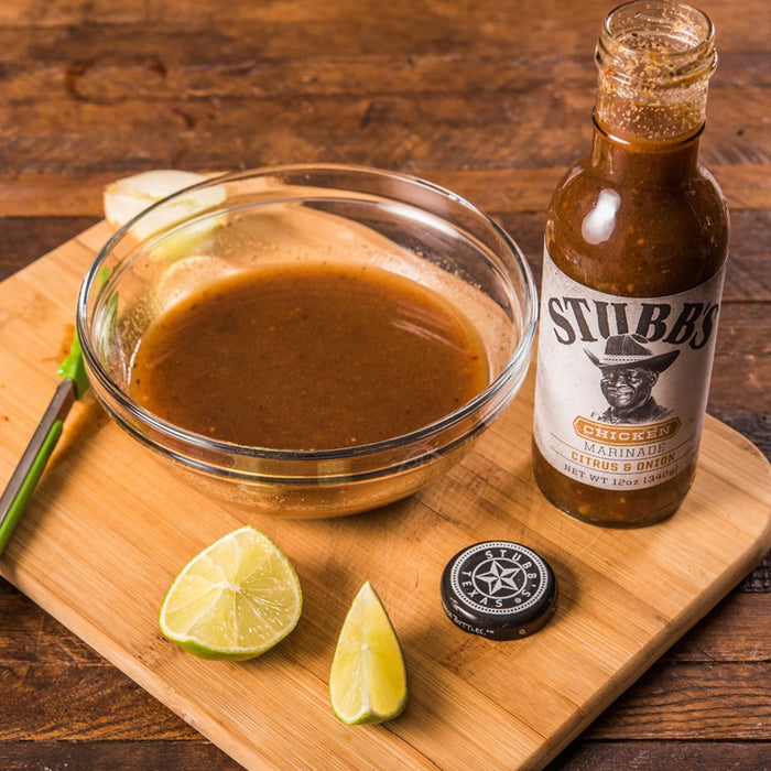 A bottle of Stubbs Chicken Marinade Citrus &amp; Onion with a bowl of marinade and lime wedges on a wooden cutting board.
