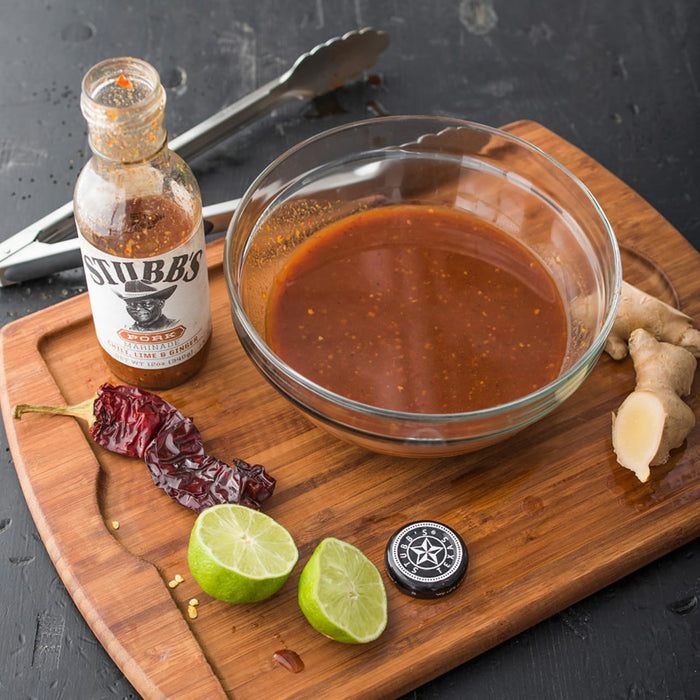 A wooden cutting board with a bowl of marinade, limes, a chili pepper, ginger, and a bottle of Stubbs Pork Marinade.