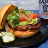 A close-up of a burger with avocado, tomato, onion, and cilantro on a bun, with a bottle of Stubbs BBQ sauce in the background.