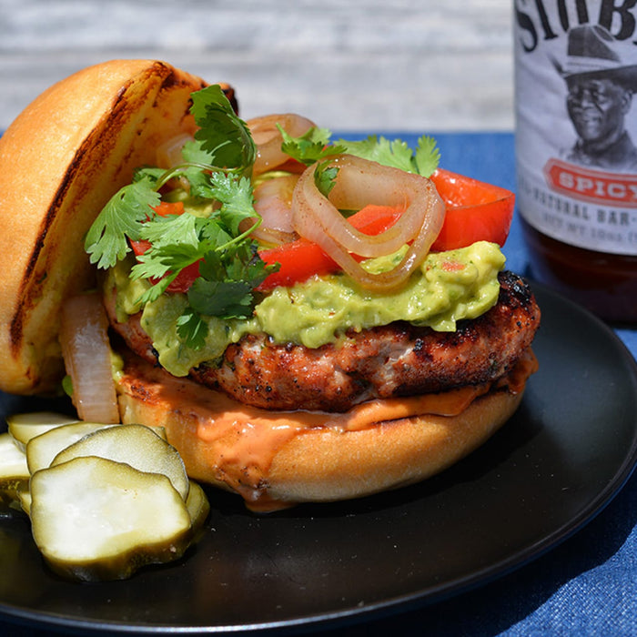 A close-up of a burger with avocado, tomato, onion, and cilantro on a bun, with a bottle of Stubbs BBQ sauce in the background.