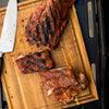 A close-up of a wooden cutting board with four pieces of grilled ribs and a silver knife.