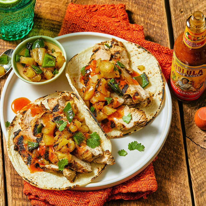 Two tacos filled with grilled chicken, pineapple, and jalapenos, with a side of salsa and a bottle of Cholula Sweet Habanero Hot Sauce.