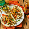 Two tacos filled with grilled chicken, pineapple, and jalapenos, with a side of salsa and a bottle of Cholula Sweet Habanero Hot Sauce.