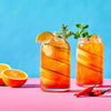 Two refreshing cocktails garnished with cucumber ribbons, mint, and lime are presented on a pink surface against a blue background alongside fresh orange halves and red chili peppers.