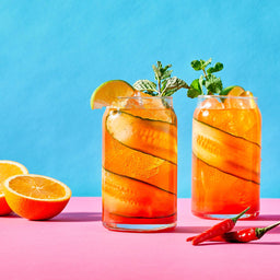 Two refreshing cocktails garnished with cucumber ribbons, mint, and lime are presented on a pink surface against a blue background alongside fresh orange halves and red chili peppers.