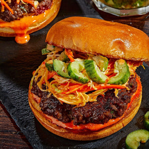 A gourmet burger featuring a seared beef patty topped with crunchy slaw and sliced cucumbers on a toasted brioche bun.