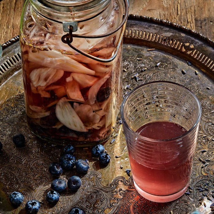 A glass jar filled with sliced onions and blueberries steeping in a reddish liquid sits on an ornate metal tray next to a glass of the infused beverage and scattered fresh blueberries.