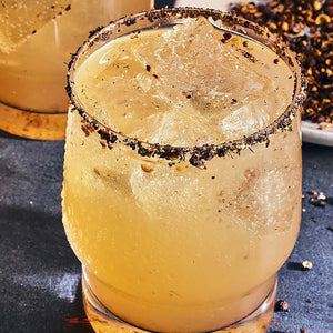 A refreshing cocktail served in a glass with a dark spiced rim and large ice cubes sits on a dark surface next to a small dish of peppercorns.