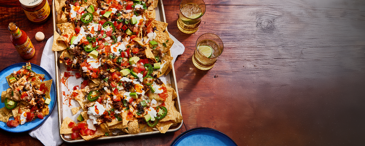 Tray of loaded nachos with various toppings on a wooden table with drinks.