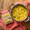 A box of Zatarains Yellow Rice sits on a red napkin next to a pot of cooked yellow rice.