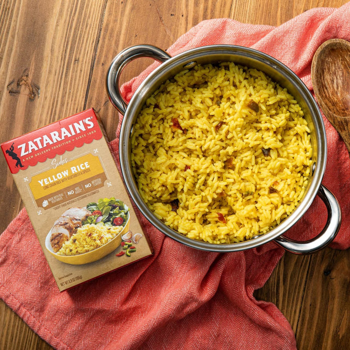 A box of Zatarains Yellow Rice sits on a red napkin next to a pot of cooked yellow rice.